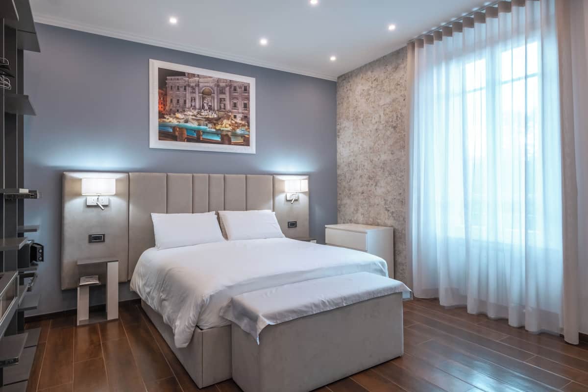 A modern hotel room with a large bed, bedside tables, a nightstand, a wall-mounted lamp, and a large window with sheer curtains.