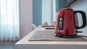 Une bouilloire rouge est posée sur une table recouverte d’un set de table, à côté d’une assiette avec une tasse, et un panier se trouve dans une pièce moderne dotée d’une fenêtre et de rideaux.