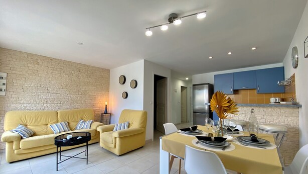 Un salon moderne avec un canapé jaune, une table à manger avec des chaises et un coin cuisine doté de meubles bleus et d’un réfrigérateur en acier inoxydable.
