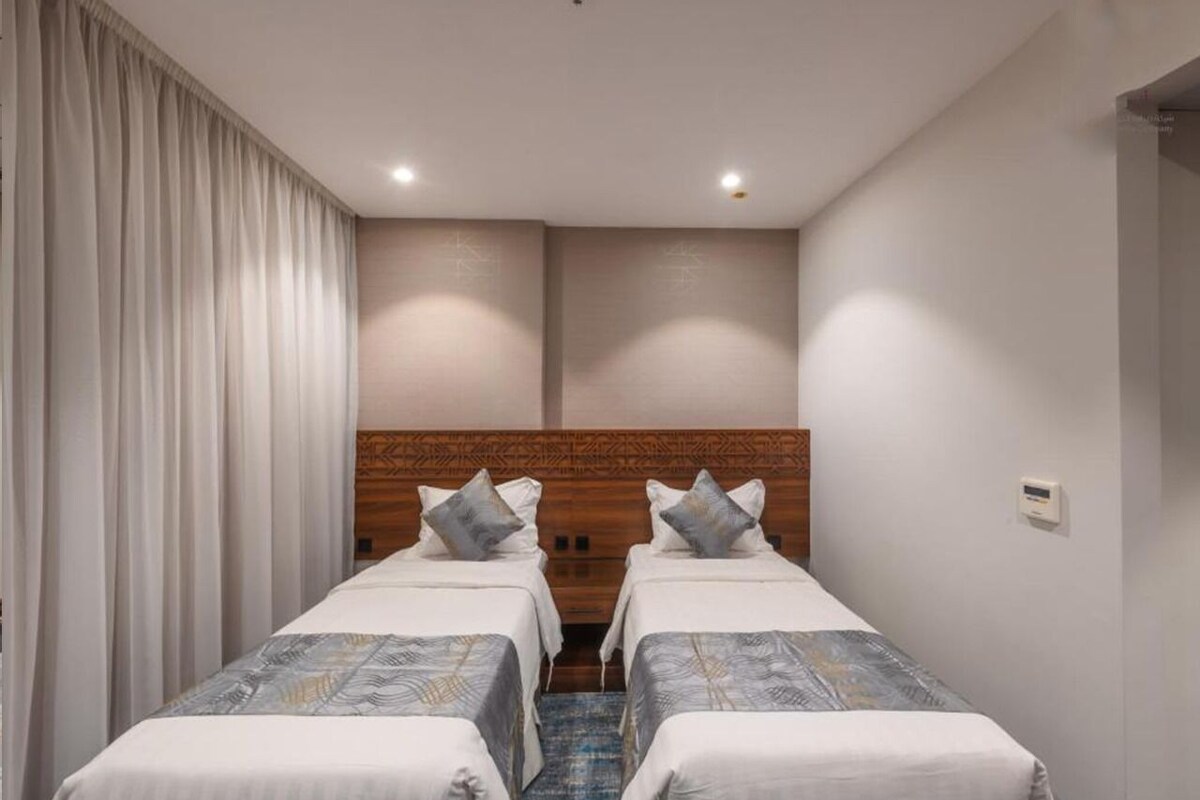A hotel room with two single beds, a wooden headboard, a white curtain, and a small wall-mounted thermostat.