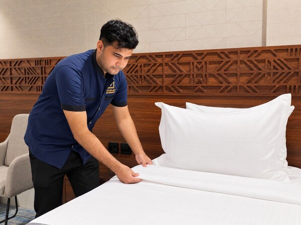 Una persona acomodando un almohadón blanco sobre una cama en una habitación con cabecera de madera y pared con diseño.