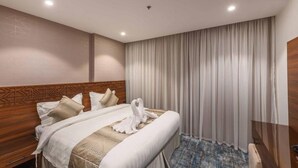 Habitación de hotel con cabecera de madera, cama con sábanas blancas, alfombra azul estampada y un amplio ventanal con cortinas blancas.