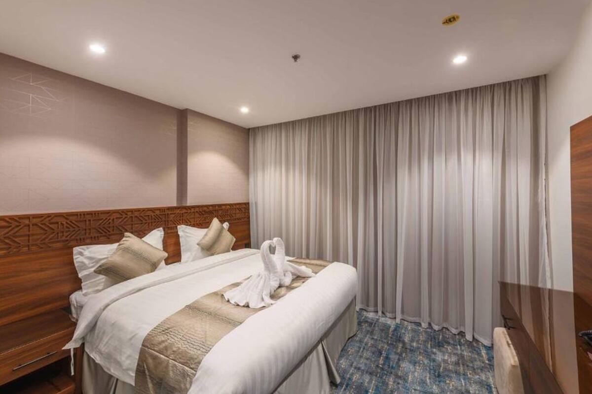 A hotel room with a wooden headboard, a bed with white linens, a blue patterned carpet, and a large window with white curtains.