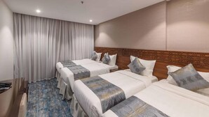 Hotelkamer met drie bedden, een houten hoofdeinde, een groot raam met gordijnen en een blauw gestreept tapijt.