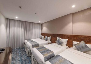 Una habitación de hotel con tres camas, un cabecero de madera, un ventanal grande con cortinas y una alfombra azul estampada.