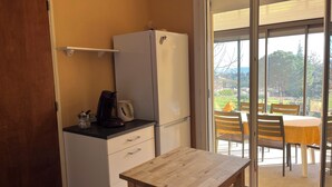Eine kompakte Küche mit Kühlschrank, Kaffeemaschine und Wasserkocher auf einer Arbeitsfläche. Vor einem Glastürschieber mit Blick auf eine Außenpatio befindet sich ein Holztisch mit Stühlen.