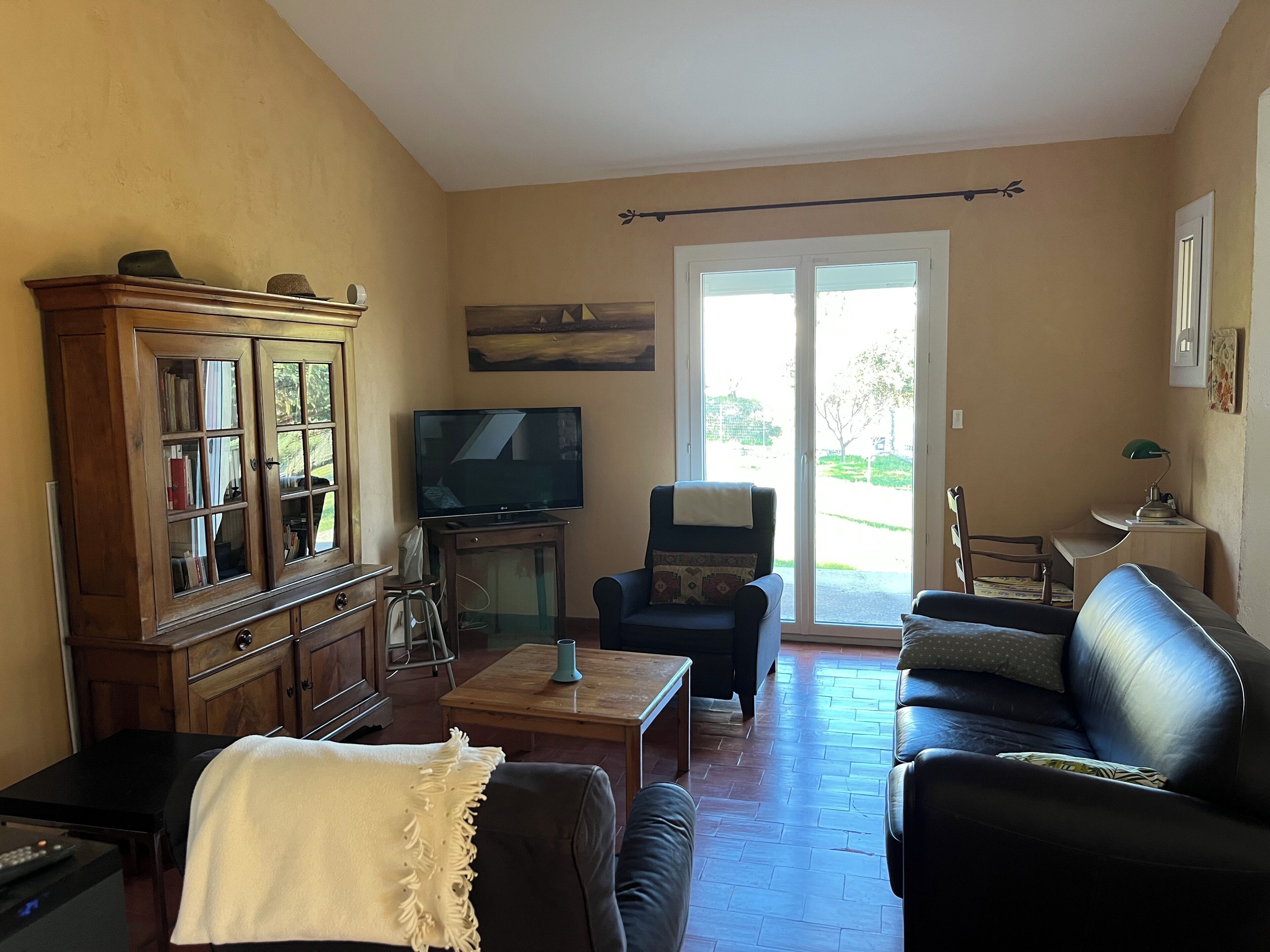 A living room with a wooden cabinet, a television, a sofa, and a chair. There is a sliding glass door leading to an outdoor area.