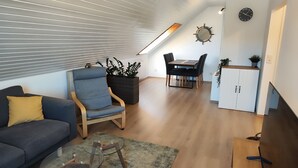 A modern living room with a sofa, a blue armchair, a glass coffee table, a dining area with a table and chairs, a white cabinet, and a skylight.