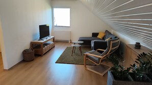 A modern living room with a flat-screen TV on a wooden stand, a sofa with cushions, a glass coffee table, and a chair. There is a window with blinds, a radiator, and a plant on the right side.
