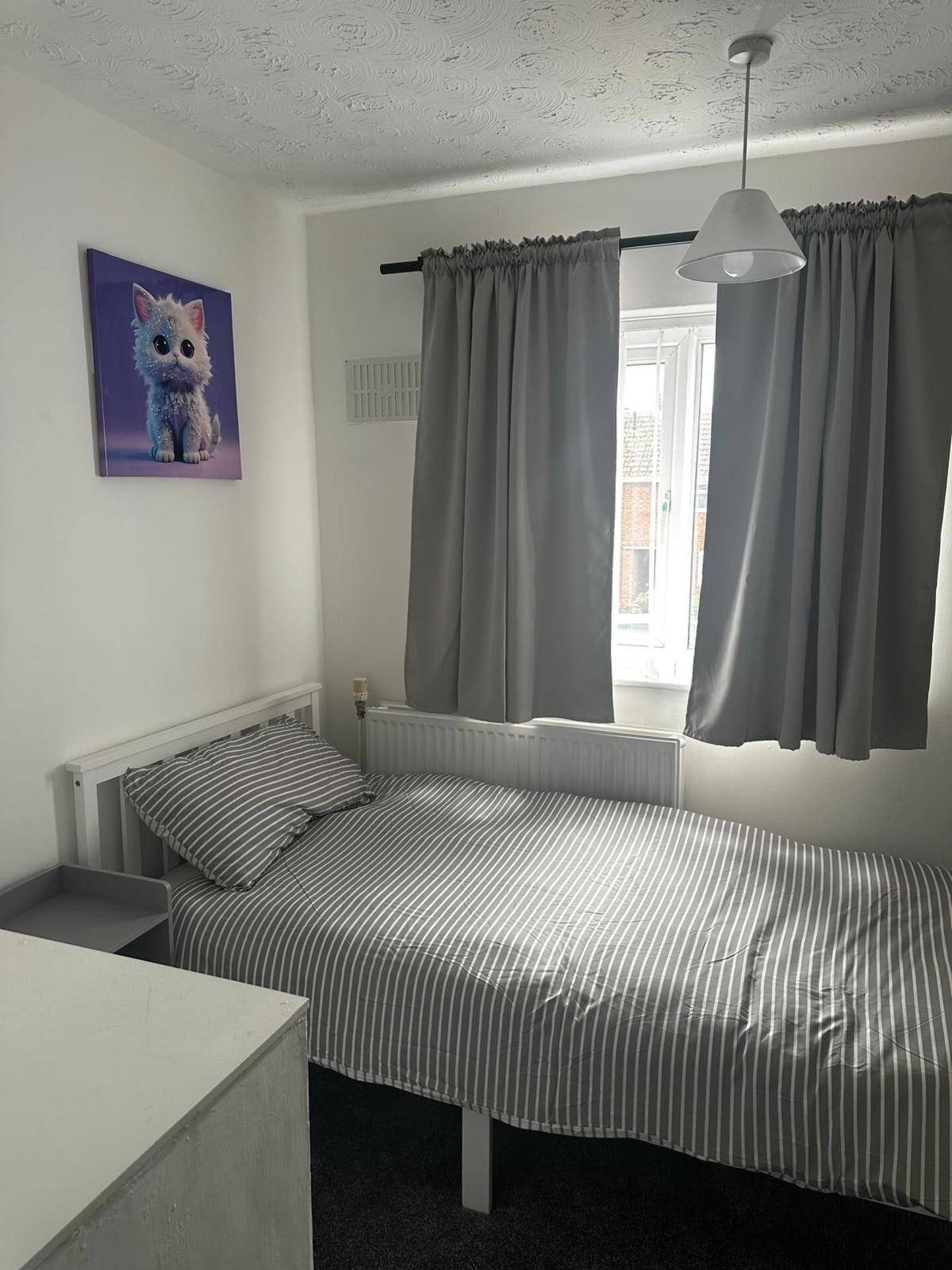 Et enkeltværelse med radiator, grå gardiner, en hængende lampe og et billede på væggen.