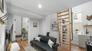 A modern living room with a sofa, a coffee table, a TV, a wooden ladder, and a kitchen area with a window.