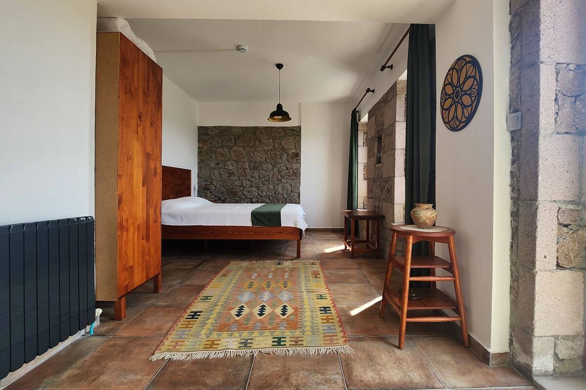 Une chambre avec un lit, une armoire en bois, un mur en pierre, une petite table avec un vase et une vue sur l’extérieur.