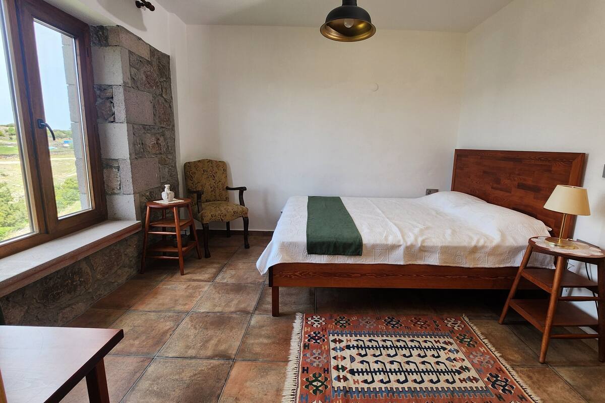 Une chambre avec un mur en pierre, un lit doté d’une tête de lit en bois, un tapis à motifs, une chaise, une petite table et une fenêtre donnant sur un paysage agréable.