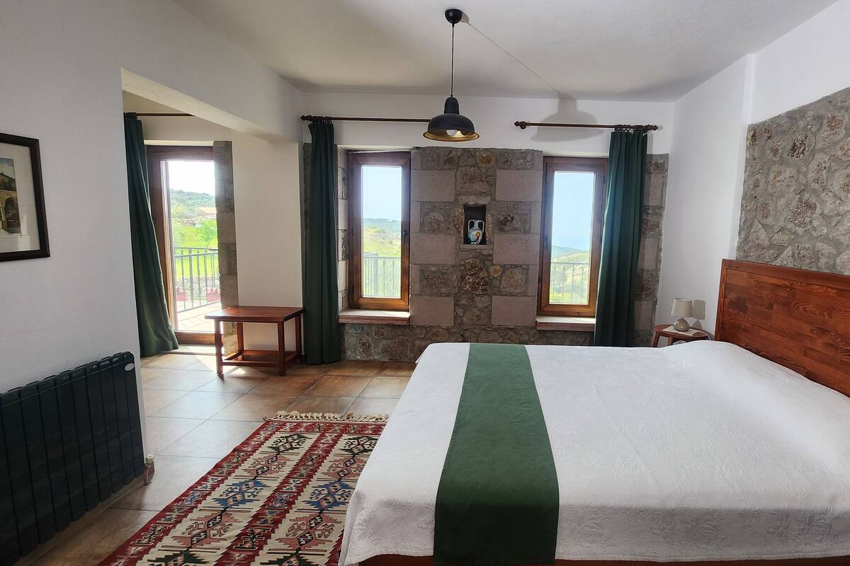 Une chambre avec un grand lit, une tête de lit en bois, un mur en pierre et une vue sur l’extérieur par une fenêtre.