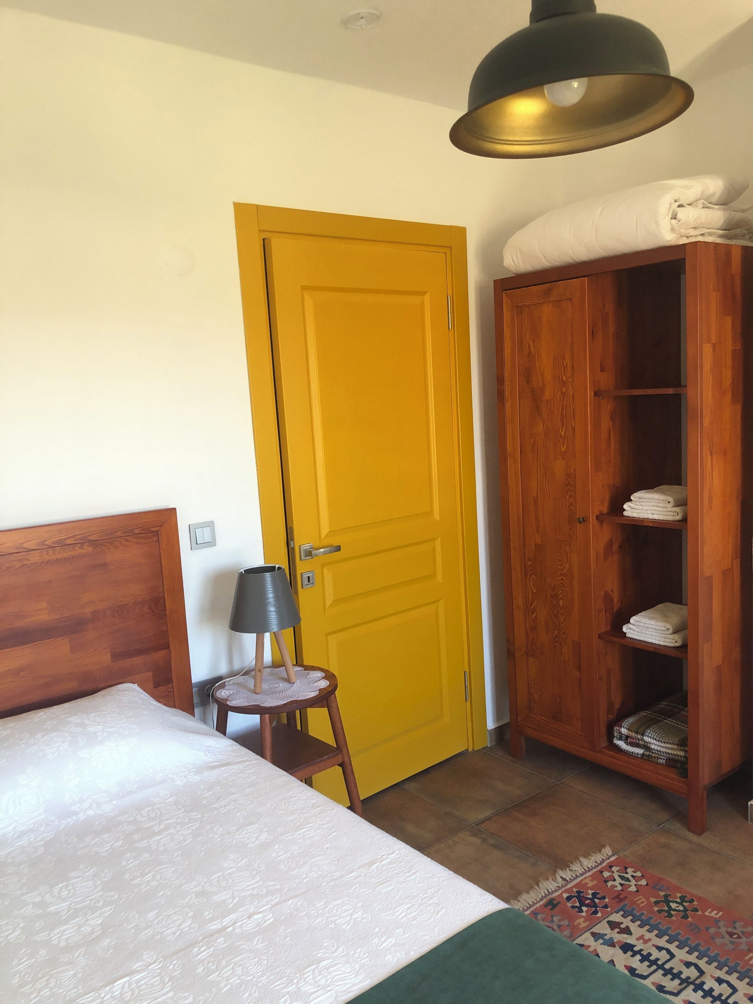 A bedroom with a wooden bed, a yellow door, a wardrobe, and a small table with a lamp.