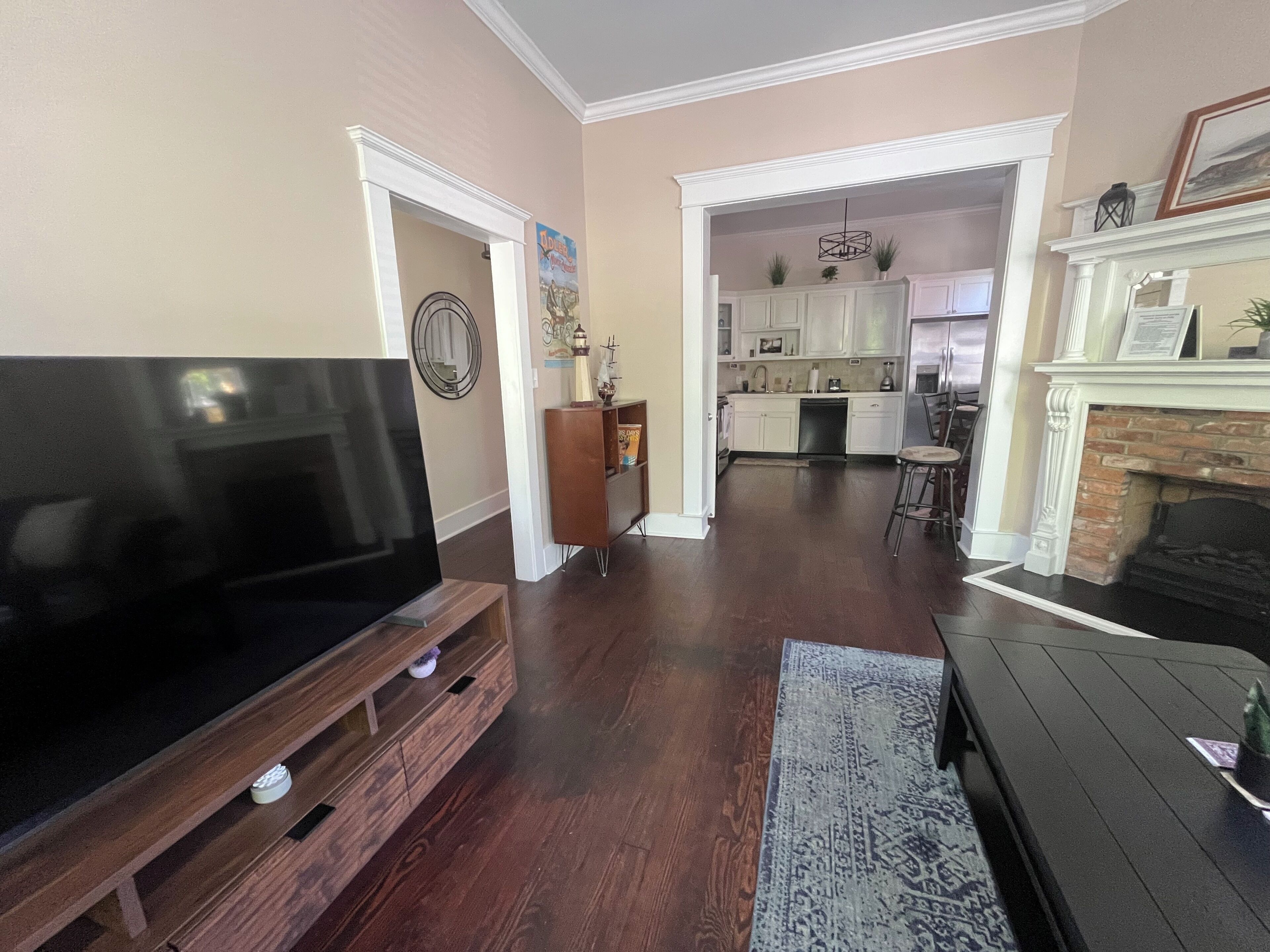 A spacious living room with a large flat-screen TV on a wooden stand, a dark-colored sofa, and a patterned rug. A kitchen with white cabinets and a brick fireplace is visible in the background.