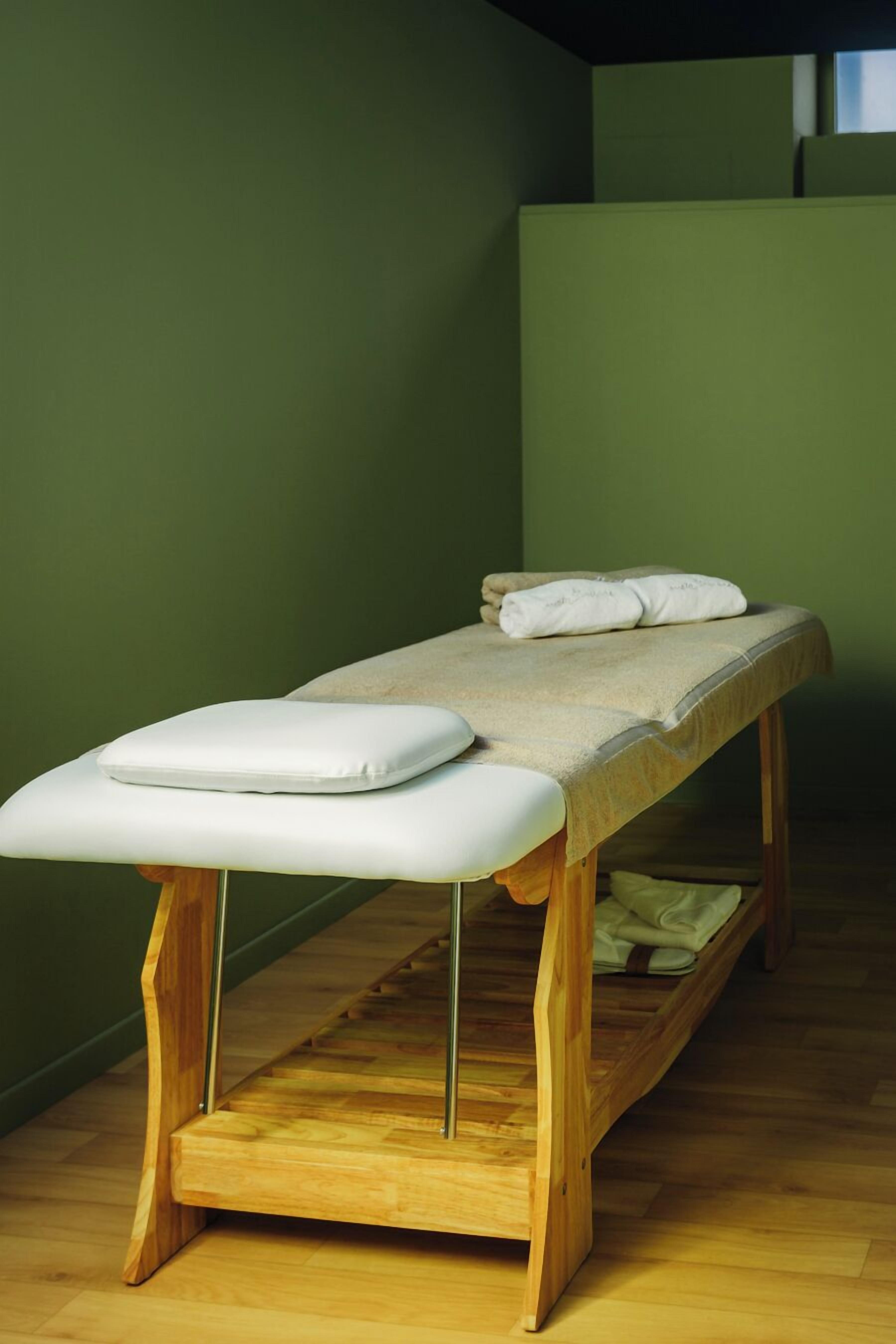 Ett massagebord i trä med en vit dyna, placerat framför en grön vägg.