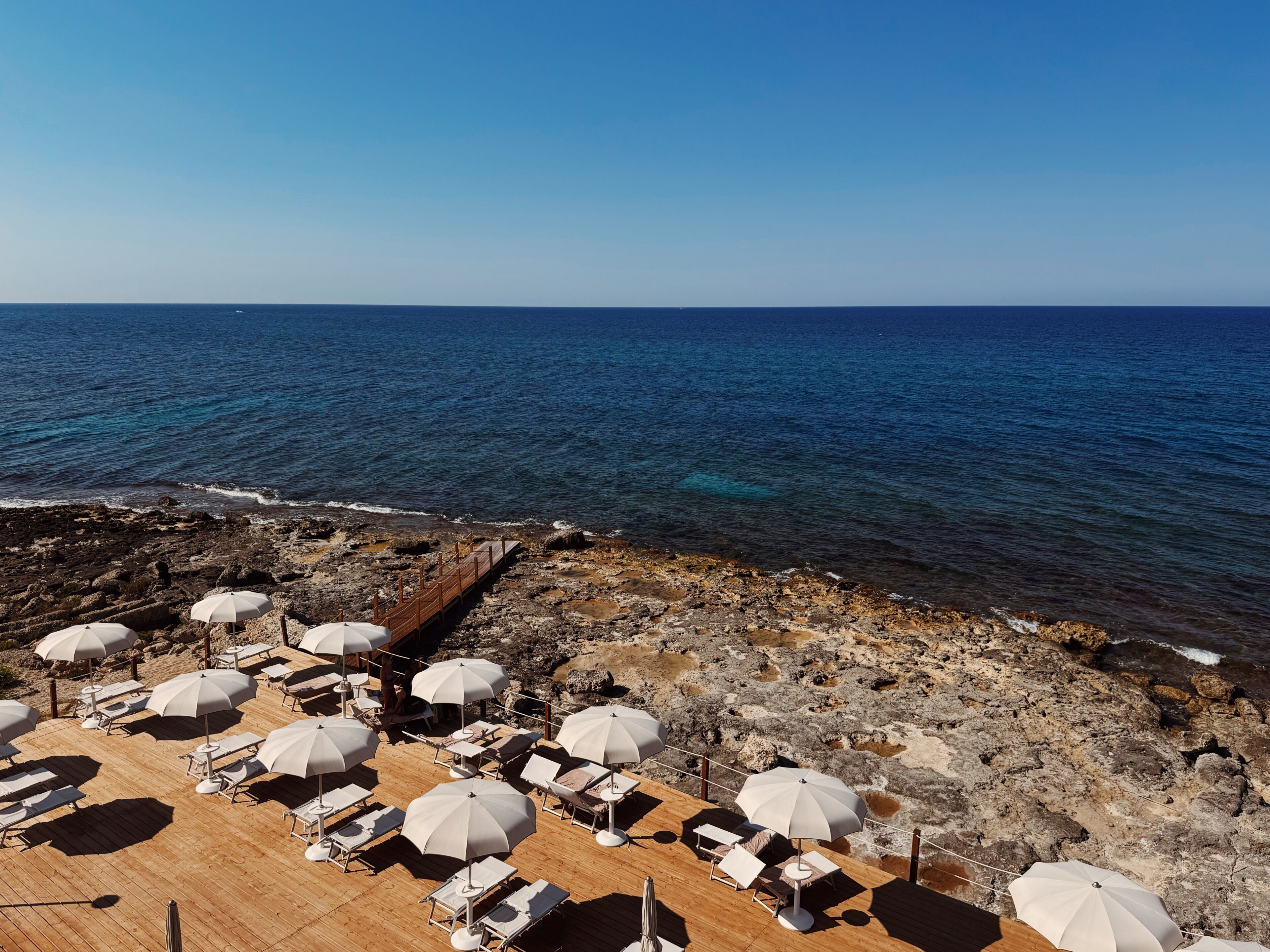 A beachside lounge area with sun loungers and umbrellas, overlooking a rocky coastline and the sea.