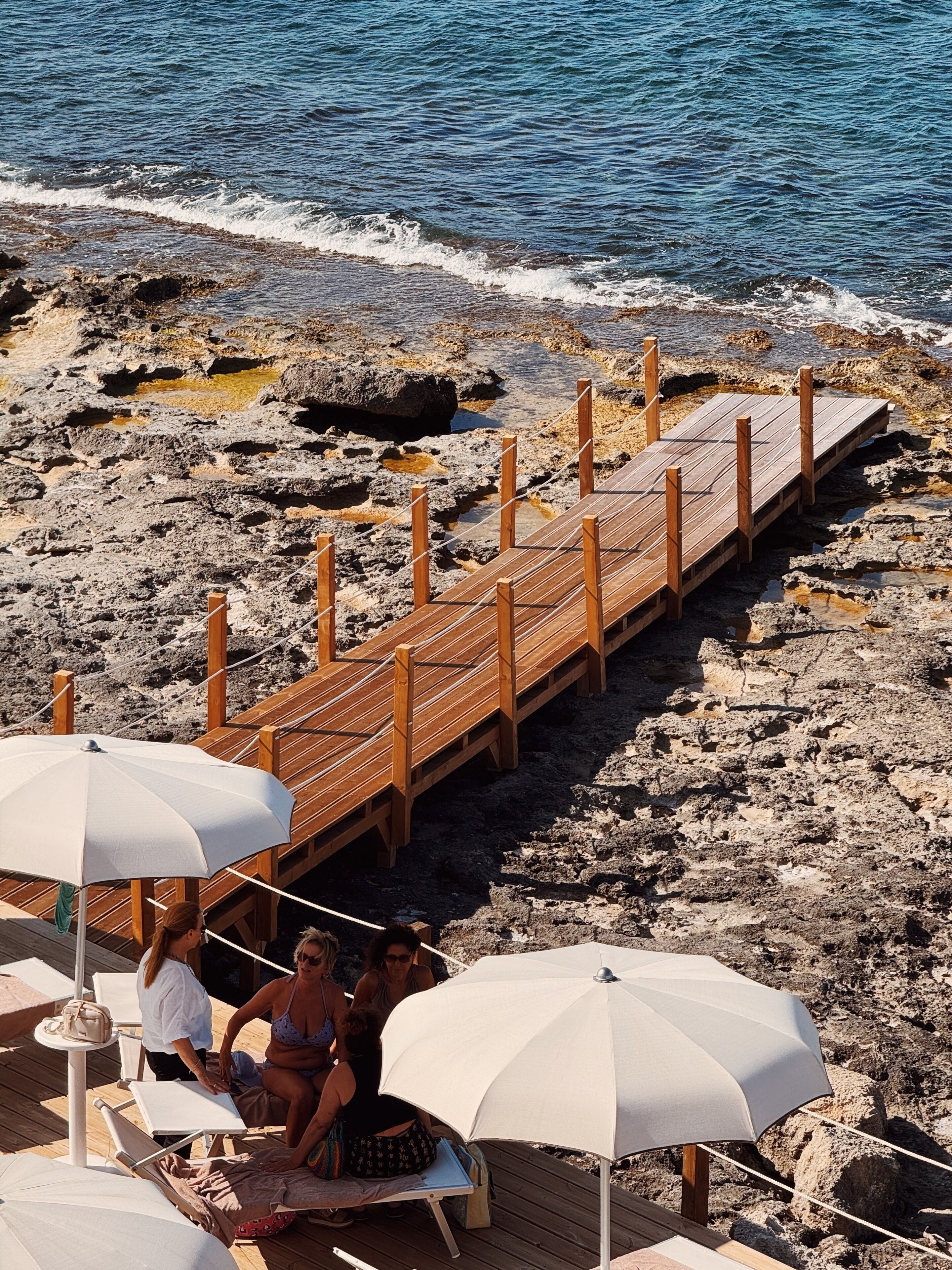 Eine Holzsteg, der zu einem Strand mit Liegestühlen und Sonnenschirmen führt.