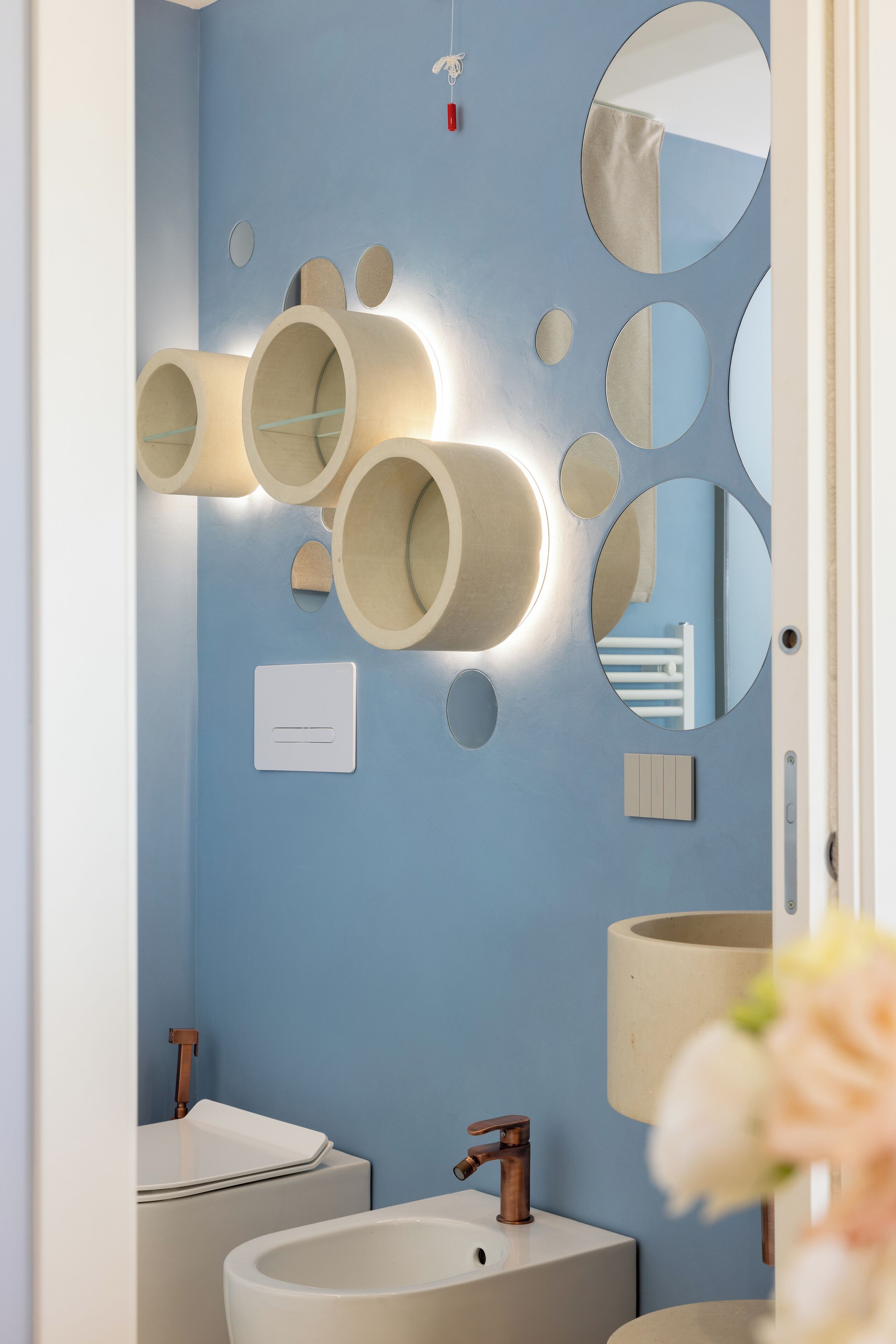 Un baño con pared azul, lavamanos blanco, espejo y lámparas cilíndricas.