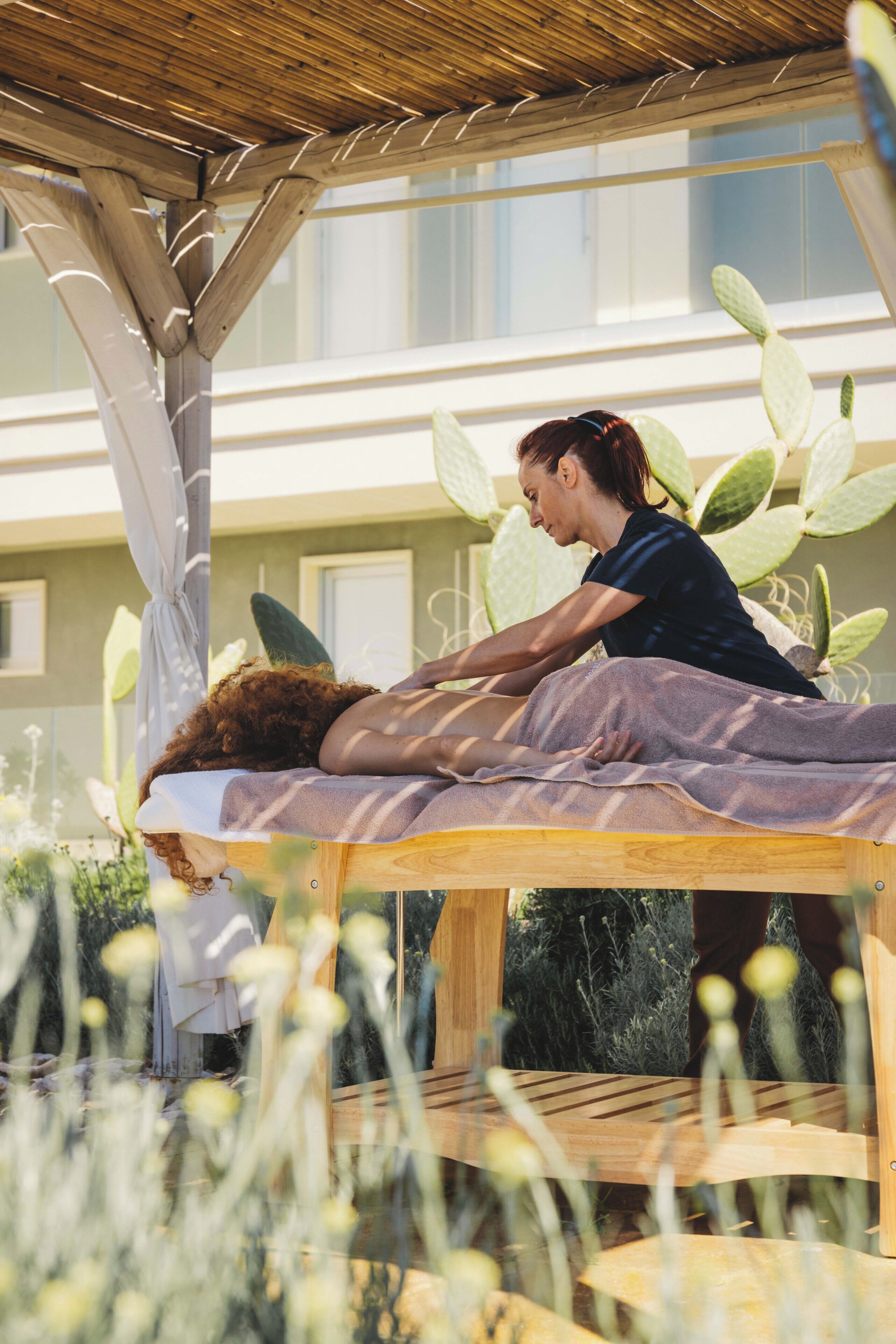 A wooden massage table with a person receiving a massage outdoors, surrounded by cacti and plants.