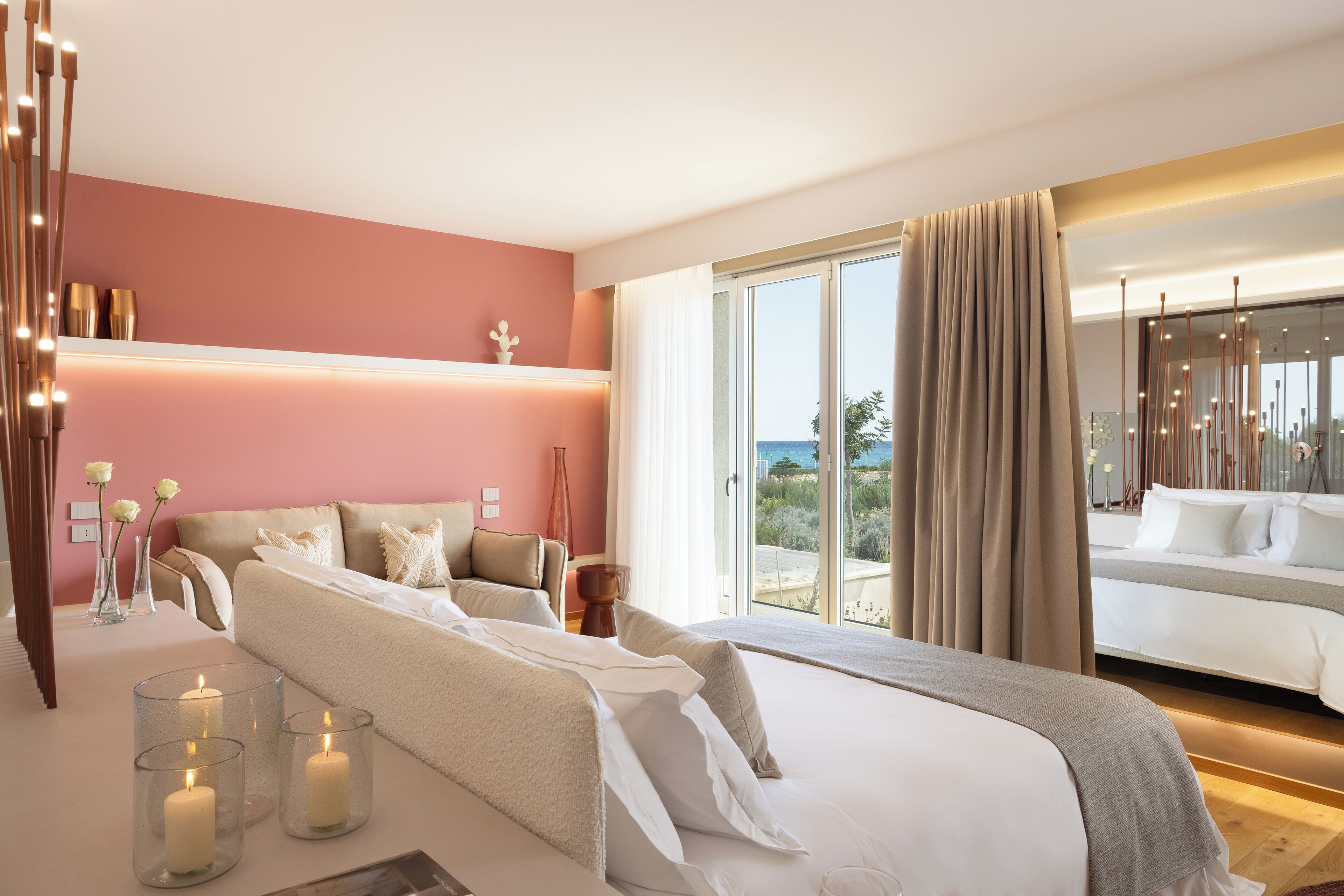 A modern hotel room with a large bed, a sofa, a dining table, and a view of the sea.