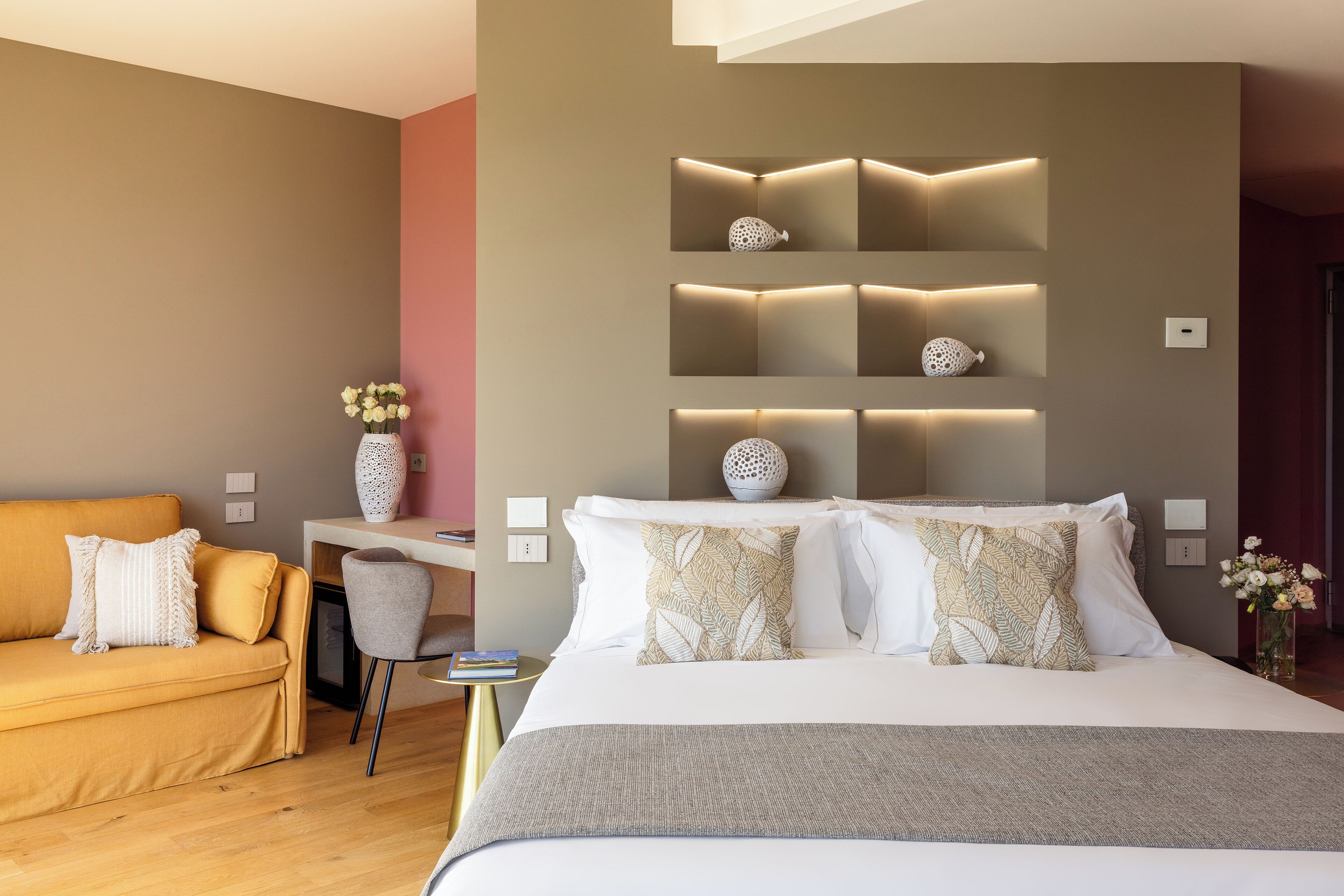 A modern hotel room with a large bed, a sofa, a desk, and a wall-mounted shelf with decorative items.