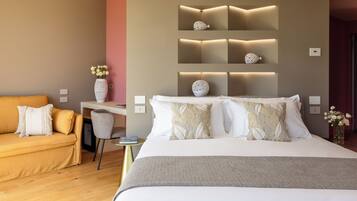 A modern hotel room with a large bed, a sofa, a desk, and a wall-mounted shelf with decorative items.