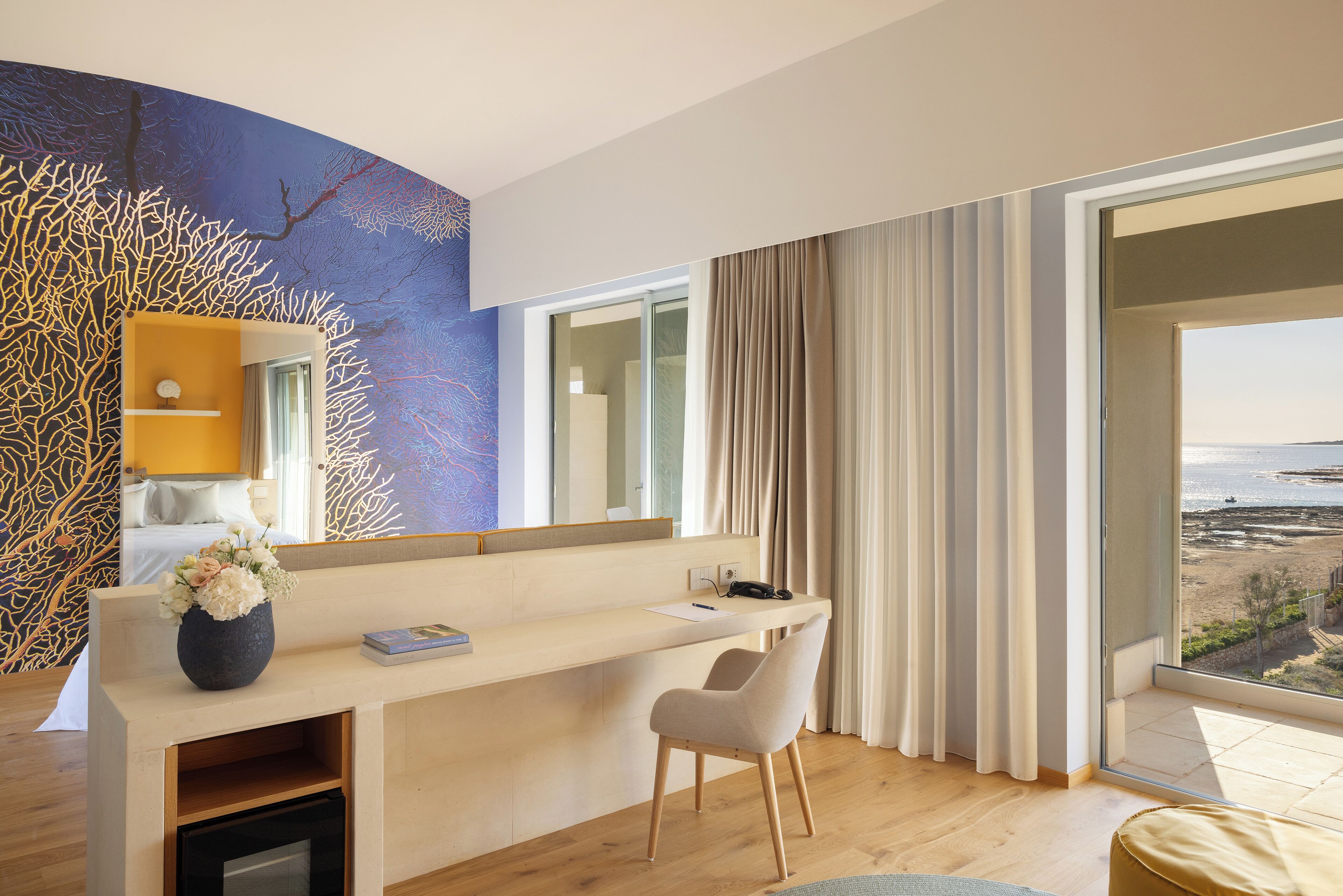 Una habitación de hotel moderna con un escritorio de madera, una silla, un florero con flores, una cama y un amplio ventanal con vista a la playa.