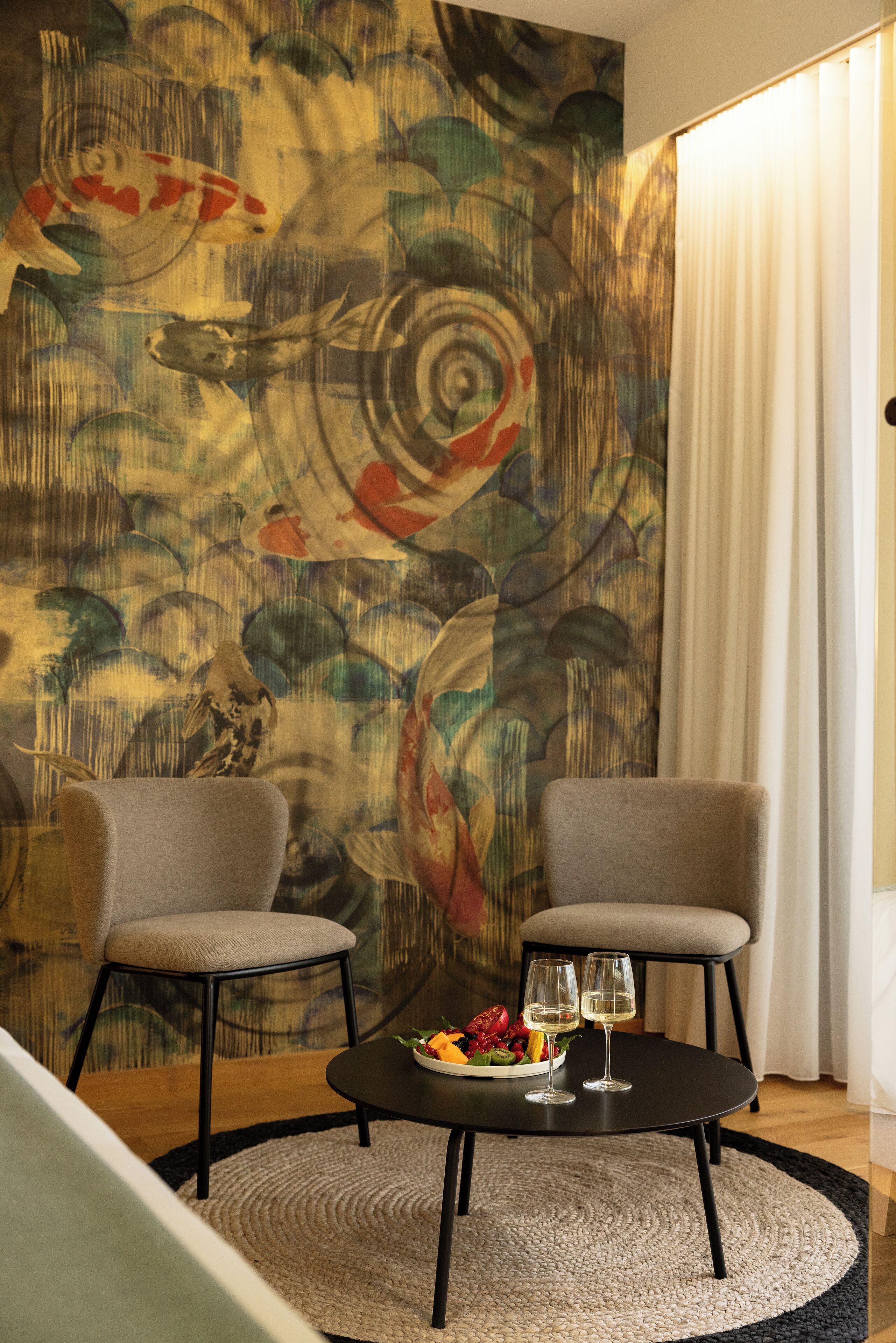 Una habitación con dos sillones, una mesa redonda con frutas y vasos, y un gran mural abstracto en la pared.
