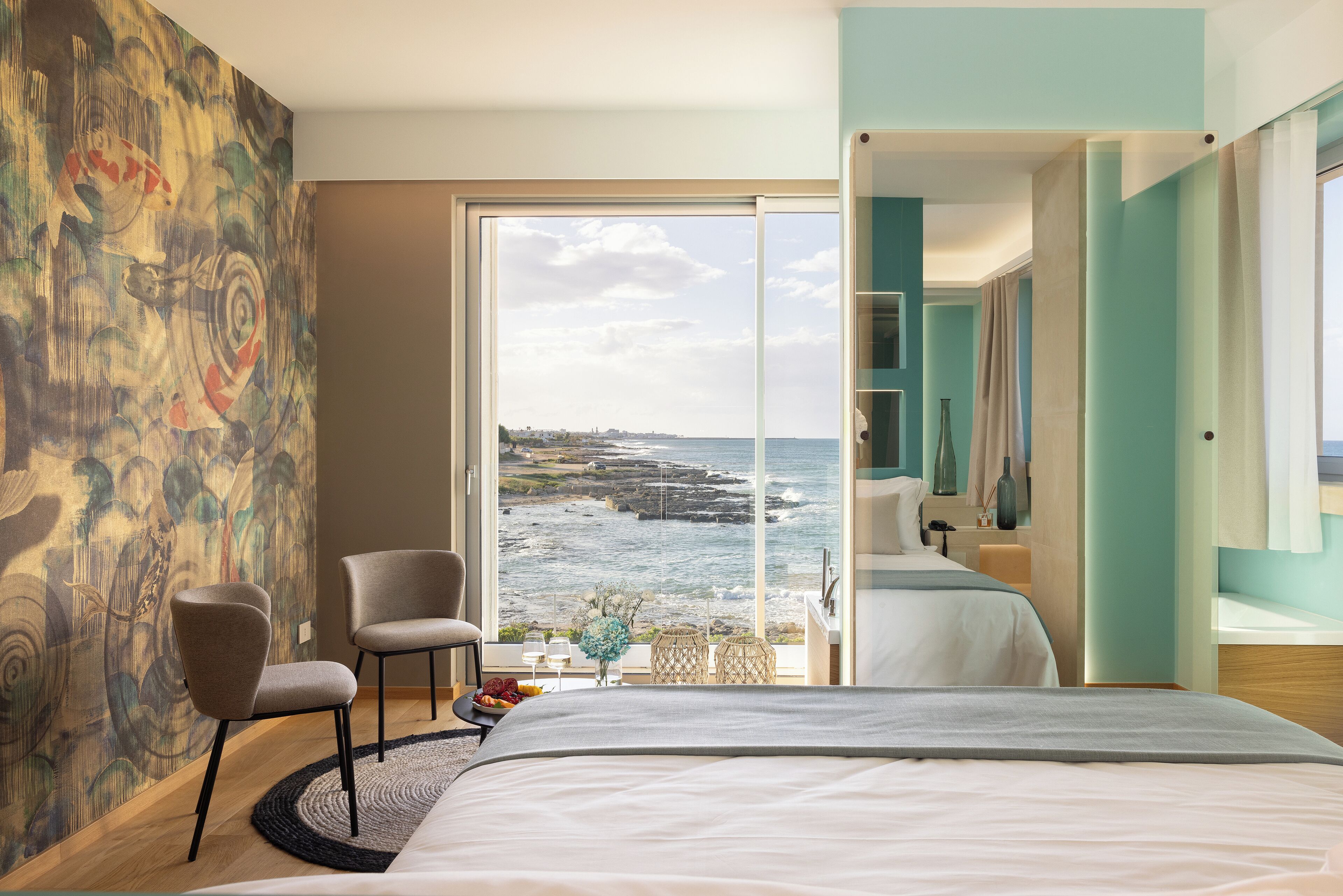 A modern hotel room with a large bed, a view of the ocean, a painting on the wall, and a seating area with a table and chairs.