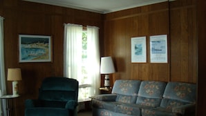 A room with wooden paneling, a blue armchair, a floral sofa, a small table with a lamp, and two framed pictures on the wall. - Spacious Grindstone Island House across from Clayton, NY (Clayton)