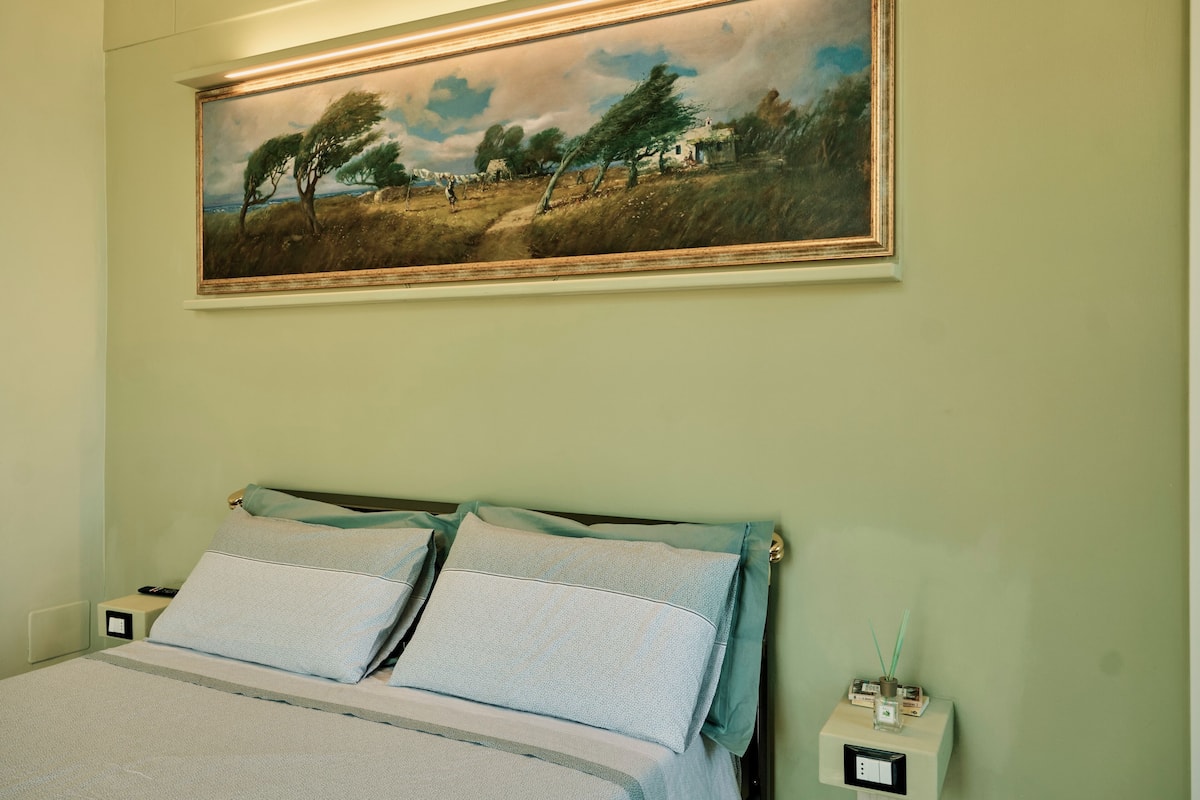 A bedroom with a bed, a nightstand, a wall-mounted painting, and a wall socket.
