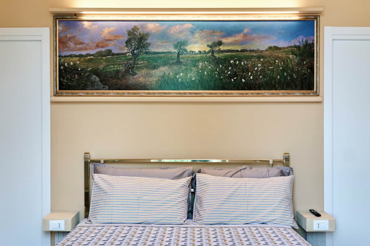 A bed with striped pillows, a painting of a landscape above it, and a remote control on a bedside table.