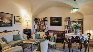 Amplia sala de estar con sofá, sillas, una mesa de centro y una mesa de comedor con sillas. Se observan estanterías llenas de libros y objetos decorativos.