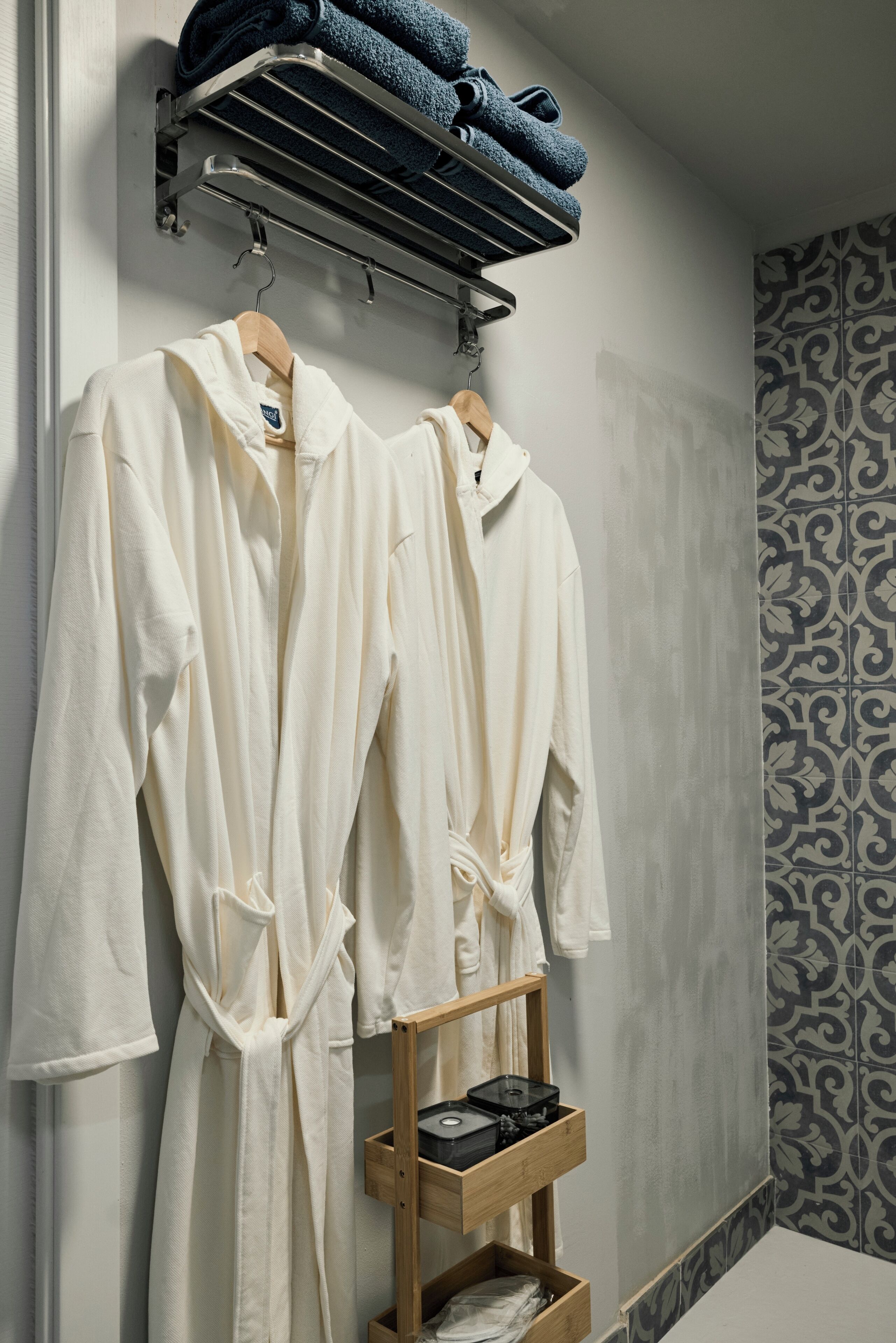 A bathroom with two white robes hanging on a rack, a towel rack with folded towels, and a wooden shelf with a tray.
