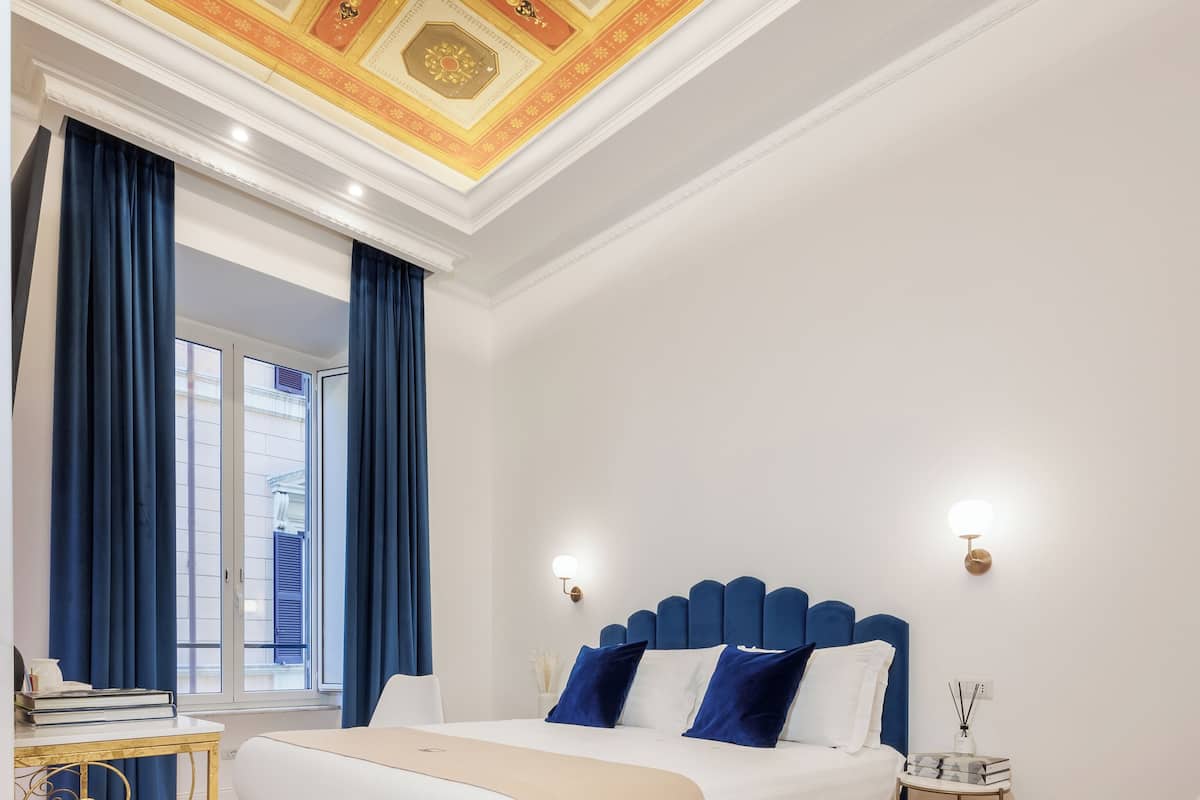 Un dormitorio con una cama grande, un escritorio, una silla, una ventana con cortinas y un techo decorativo.