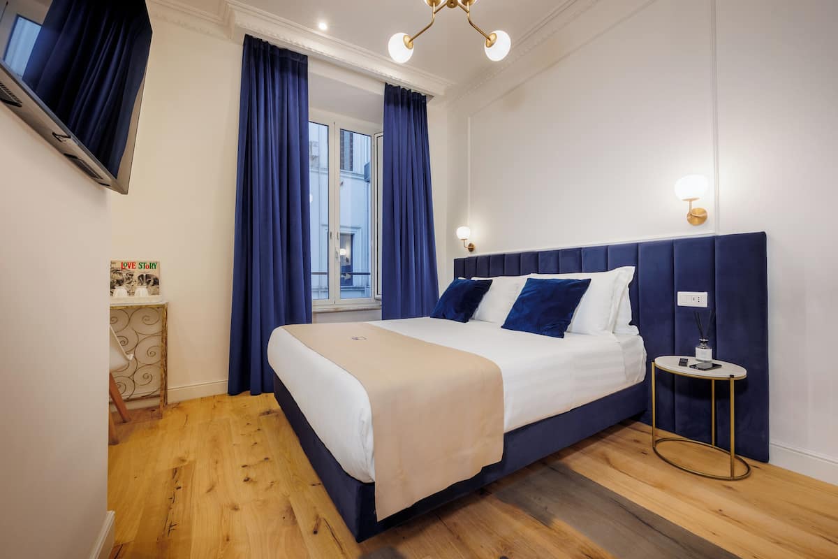 Un dormitorio moderno con una cama grande, una mesita de noche, un televisor montado en la pared y una ventana con cortinas.