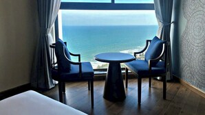 A room with a large window overlooking the ocean, two chairs, a small round table, and a bed. - Apec Mandala Mui Ne - Sapphire Apartment (Mui Ne)