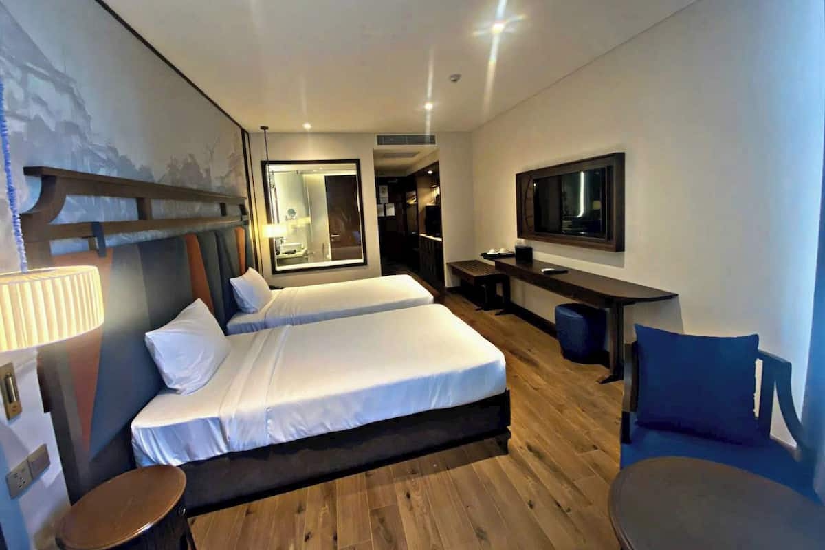 A hotel room with two beds, a desk, a chair, a TV, and a bathroom visible through an open door.