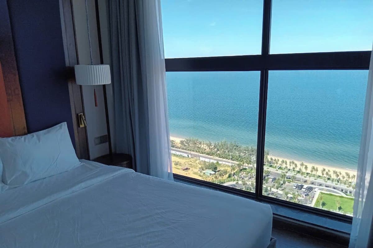 A hotel room with a large bed, a view of the ocean, and a beach visible through the window.