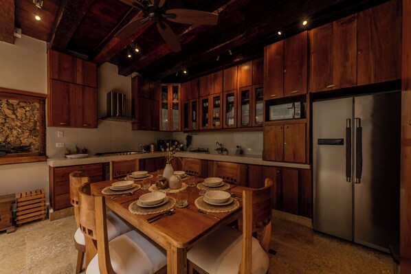 Una mesa de comedor de madera dispuesta con platos, tazones y cubiertos, rodeada de sillas de madera. Se observa un refrigerador de acero inoxidable a la derecha.