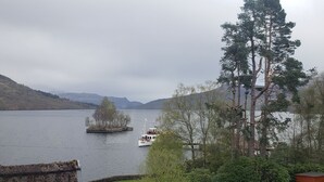 A lakeside view with a boat docked near a small island, surrounded by trees and hills. - Stronachlachar Lodge - spacious with cinematic views, direct access to loch. (Stirling)