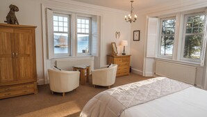 A bedroom with a large bed, two chairs, a wooden dresser, a chest of drawers, a chandelier, and a view of the outdoors through the windows. - Stronachlachar Lodge - spacious with cinematic views, direct access to loch. (Stirling)