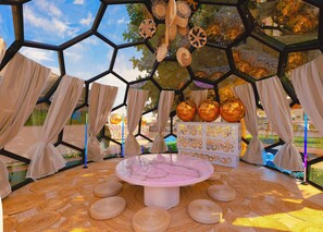 A hexagonal glass structure with woven mats on the floor, a central table, and decorative woven items hanging from the ceiling.