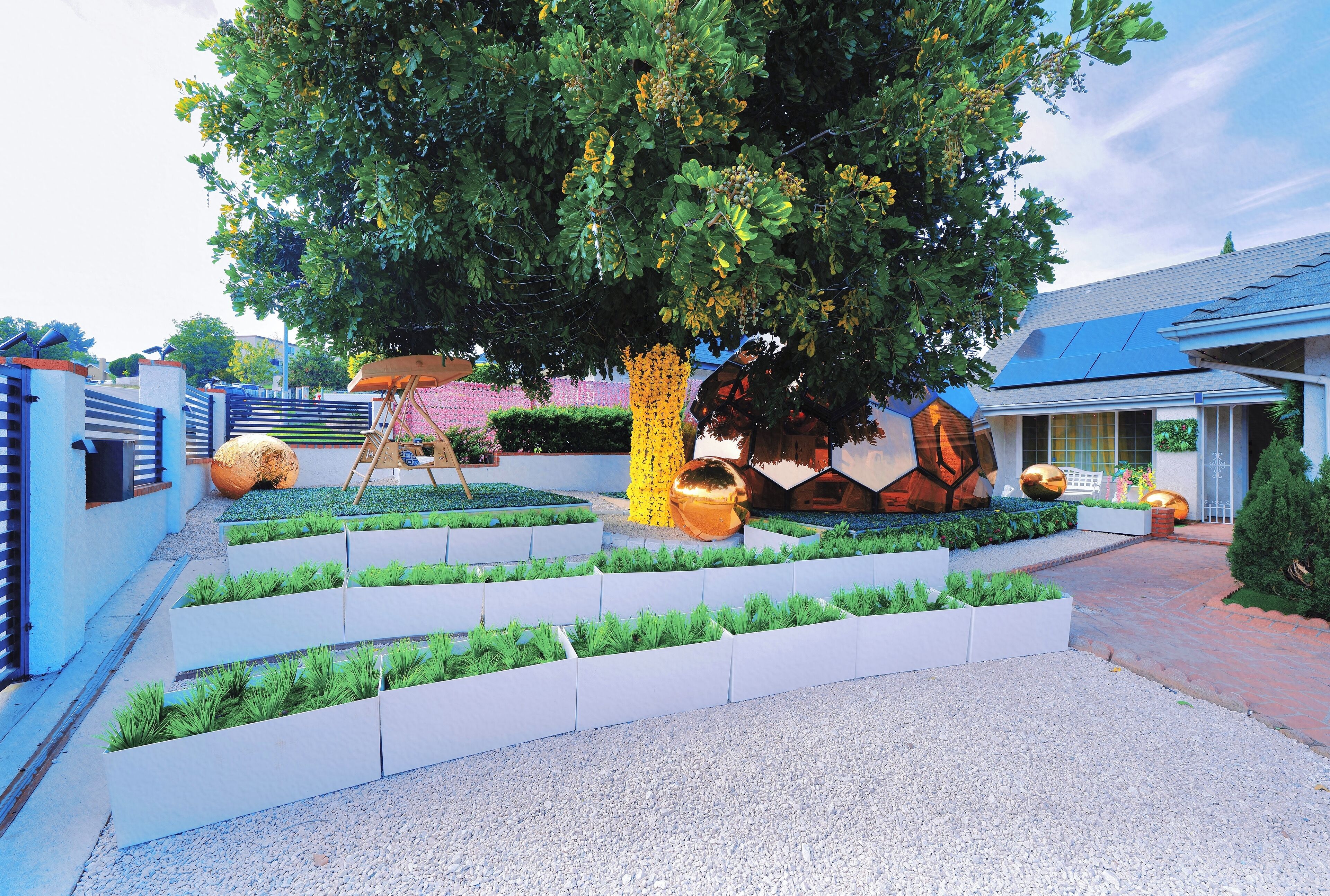 A landscaped garden with a large tree, white planters, and a pathway leading to a building with a blue roof.