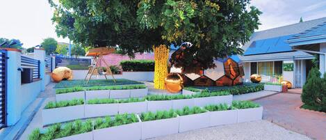 A landscaped garden with a large tree, white planters, and a pathway leading to a building with a blue roof.