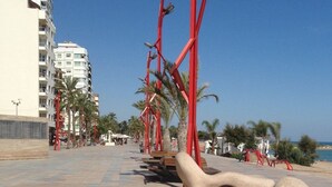 A promenade with red metal structures, palm trees, and a building with balconies. - Lovely Holiday Place with pool, A/C, TV, WIFI - Playa Sol Vinaros (VC)