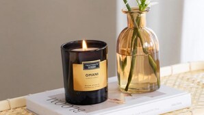 A black candle labeled "OMANI" on a white book, a clear glass vase with green plants, and a wicker basket. - Villa El Dorado B&B and Wellness (Competa)