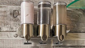 A stainless steel water dispenser with a transparent lid, mounted on a wooden wall.